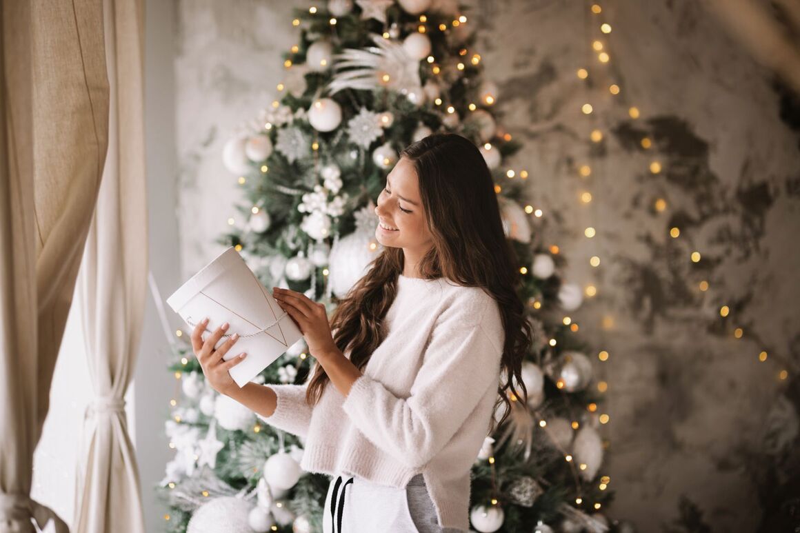 Schöne dunkelhaarige Frau in weißem Pullover und Hosen gekleidet steht neben dem Fenster auf dem Hintergrund der Weihnachtsbaum und hält ein Geschenk